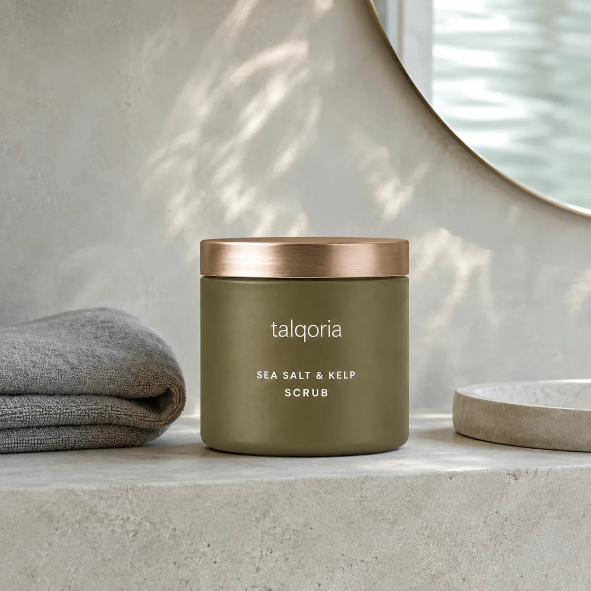 A matte olive green jar of talqoria Sea Salt & Kelp Scrub with a metallic rose gold lid, styled on a light concrete surface next to a folded gray towel and a round mirror.
