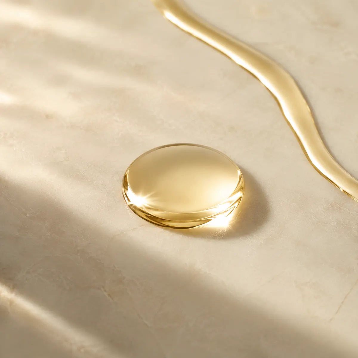 A close-up of a glossy, translucent golden liquid droplet resting on a light beige stone surface, with a matching curved streak of the same golden liquid in the upper right.