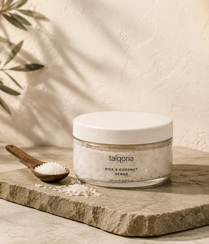 A jar of talqoria Rice & Coconut Scrub sits on a stone slab next to a wooden spoon filled with the scrub, set against a textured beige wall with soft leaf shadows.