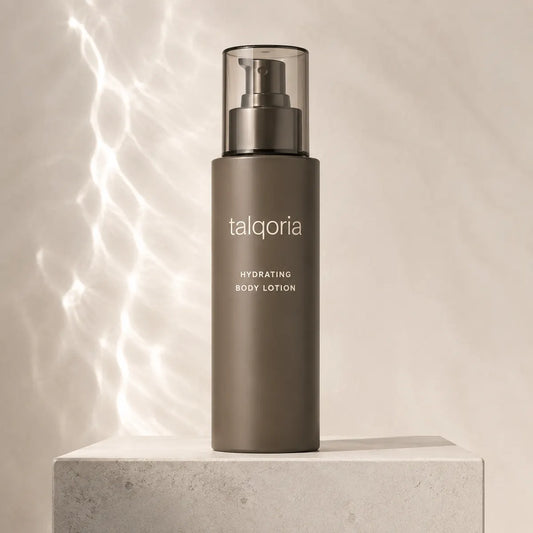 A tall matte taupe body lotion bottle with a clear cap and pump, standing on a light stone pedestal against a warm beige background with soft light streaks.