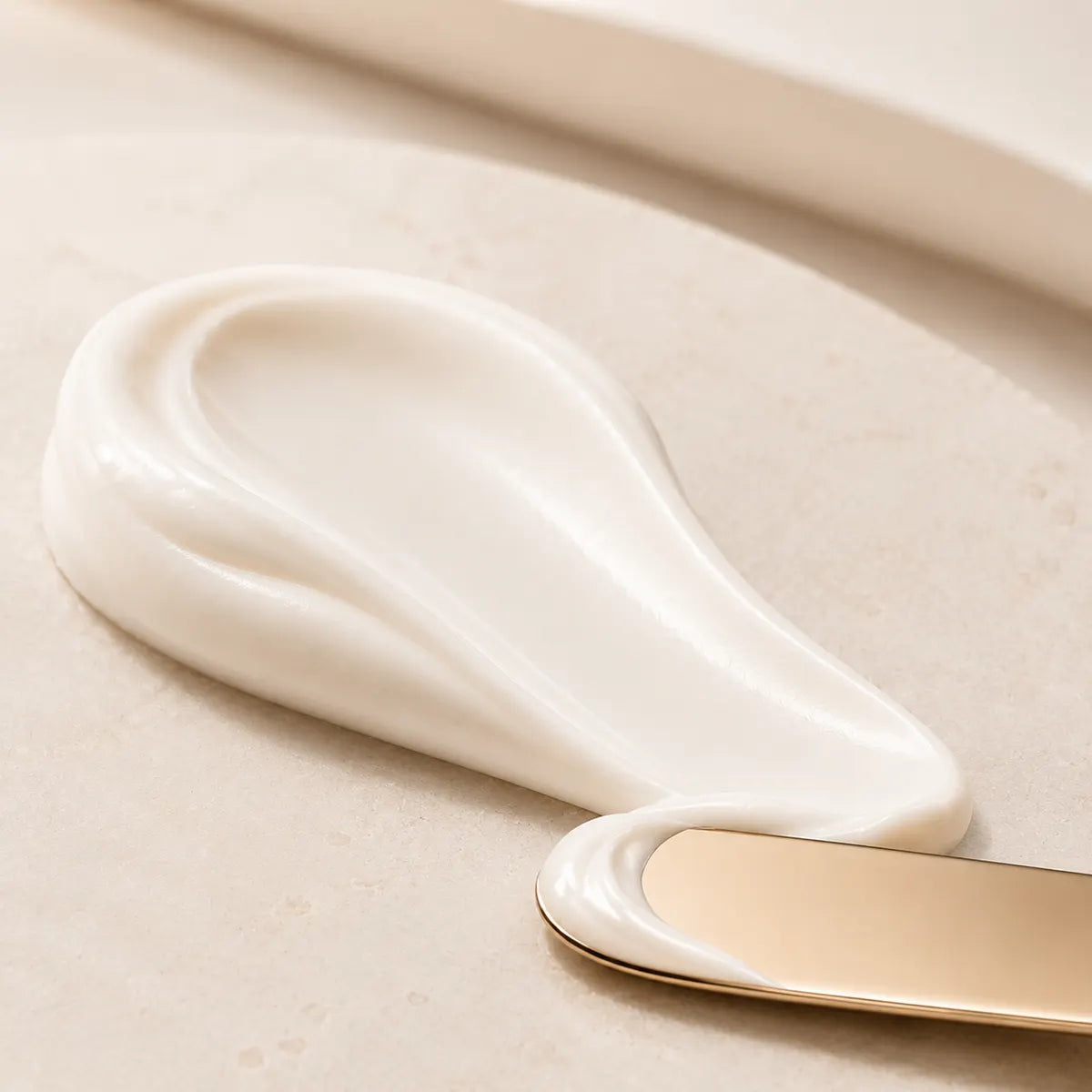 A close-up of a thick, creamy off-white cosmetic or skincare product swirled on a light beige surface, with a gold-handled spatula partially visible at the bottom right.