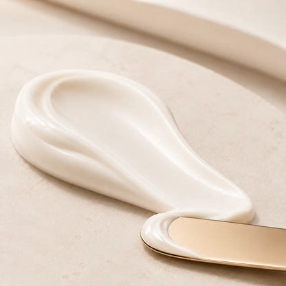 A close-up of a thick, creamy off-white cosmetic or skincare product swirled on a light beige surface, with a gold-handled spatula partially visible at the bottom right.