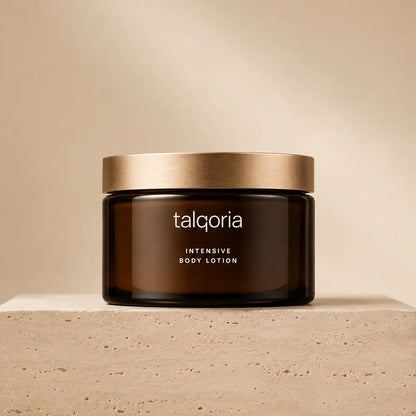 A dark amber glass jar of talqoria Intensive Body Lotion with a brushed gold lid, displayed on a textured beige stone pedestal against a matching beige background.