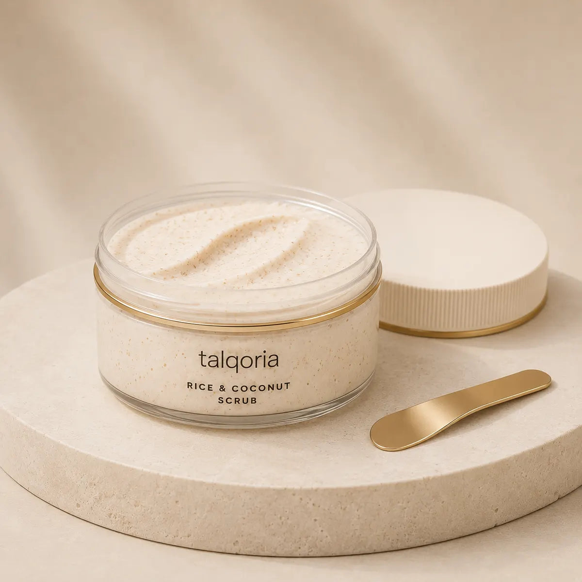 A clear jar of talqoria Rice & Coconut Scrub sits open on a round stone pedestal, with the white lid resting beside it and a small gold spatula on a beige fabric backdrop.