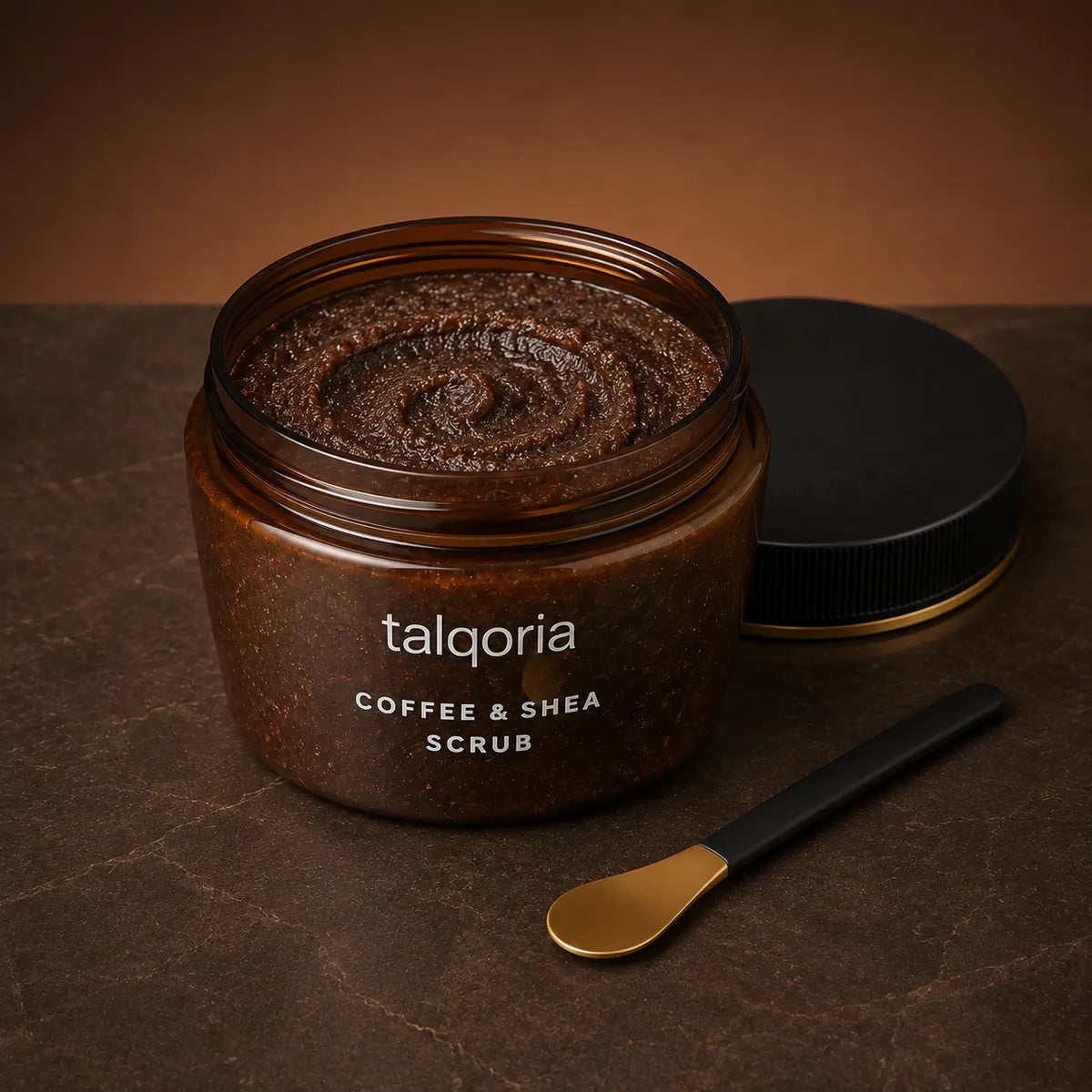 An open amber glass jar of talqoria Coffee & Shea Scrub, showing the thick dark brown scrub inside, with its black lid resting beside it on a dark brown textured surface. A small black-handled spoon with a gold-colored bowl is placed in front.