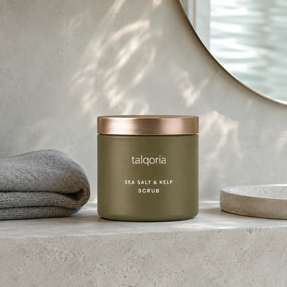 A matte olive green jar of talqoria Sea Salt & Kelp Scrub with a metallic rose gold lid, styled on a light concrete surface next to a folded gray towel and a round mirror.