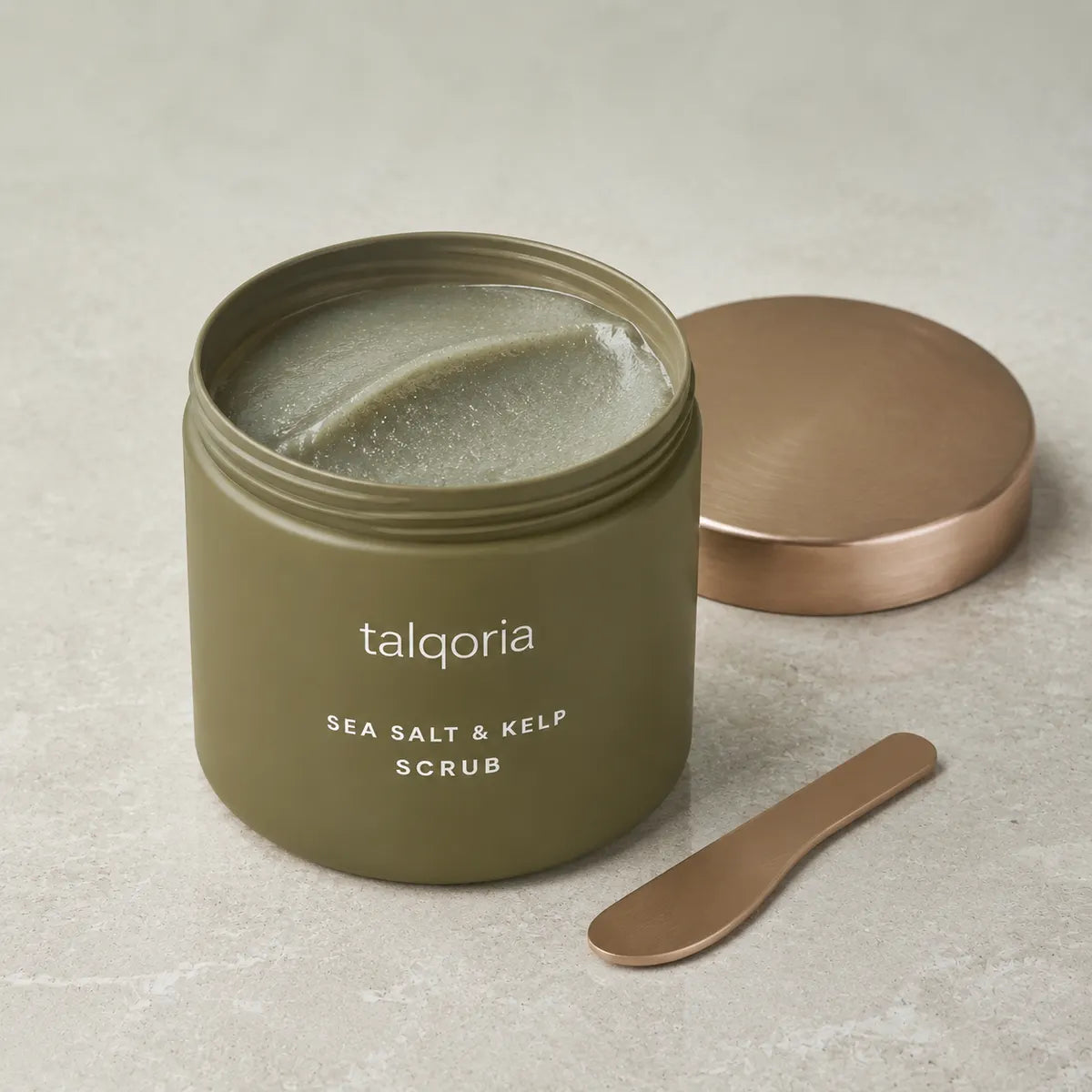 An open olive green jar of talqoria Sea Salt & Kelp Scrub, showing the textured scrub inside, with its metallic rose gold lid resting beside it on a light gray stone surface.