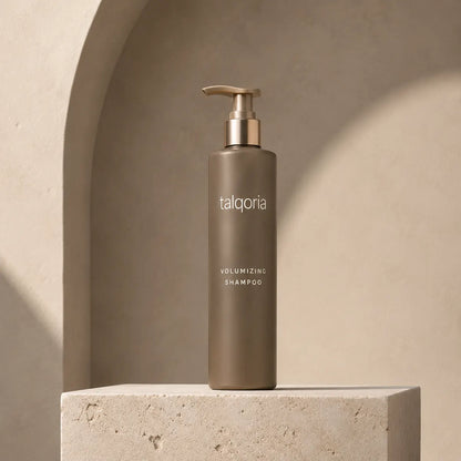 A tall taupe pump bottle of talqoria Volumizing Shampoo stands on a textured stone pedestal against a warm beige arched backdrop.