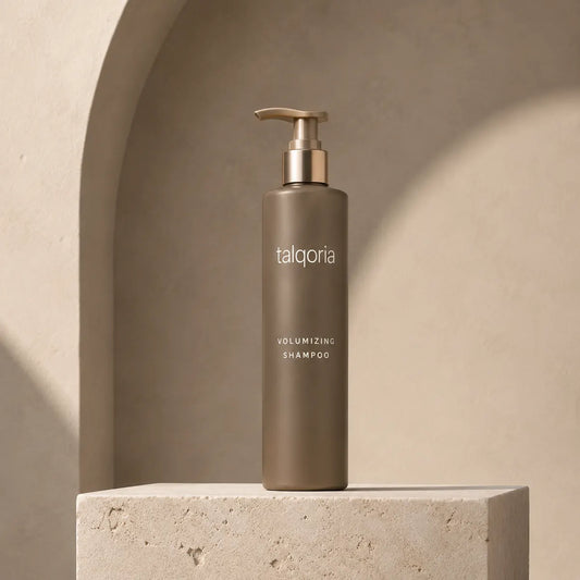 A tall taupe pump bottle of talqoria Volumizing Shampoo stands on a textured stone pedestal against a warm beige arched backdrop.