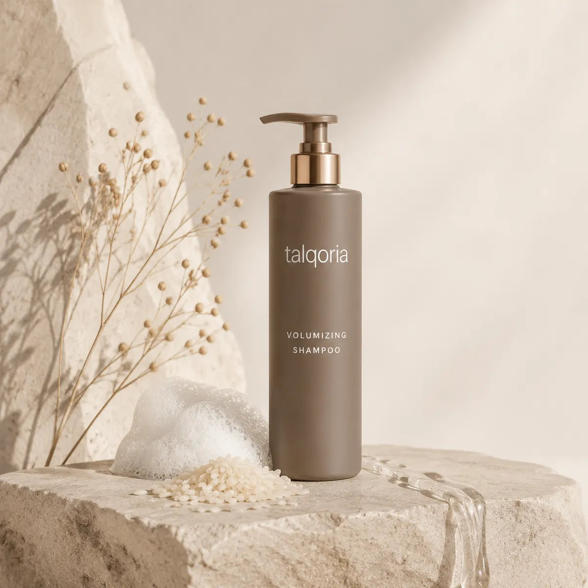 A bottle of talqoria Volumizing Shampoo in a matte taupe-brown color with a gold pump dispenser, styled on a textured stone pedestal with dried flowers and a dollop of white foam against a warm beige background.