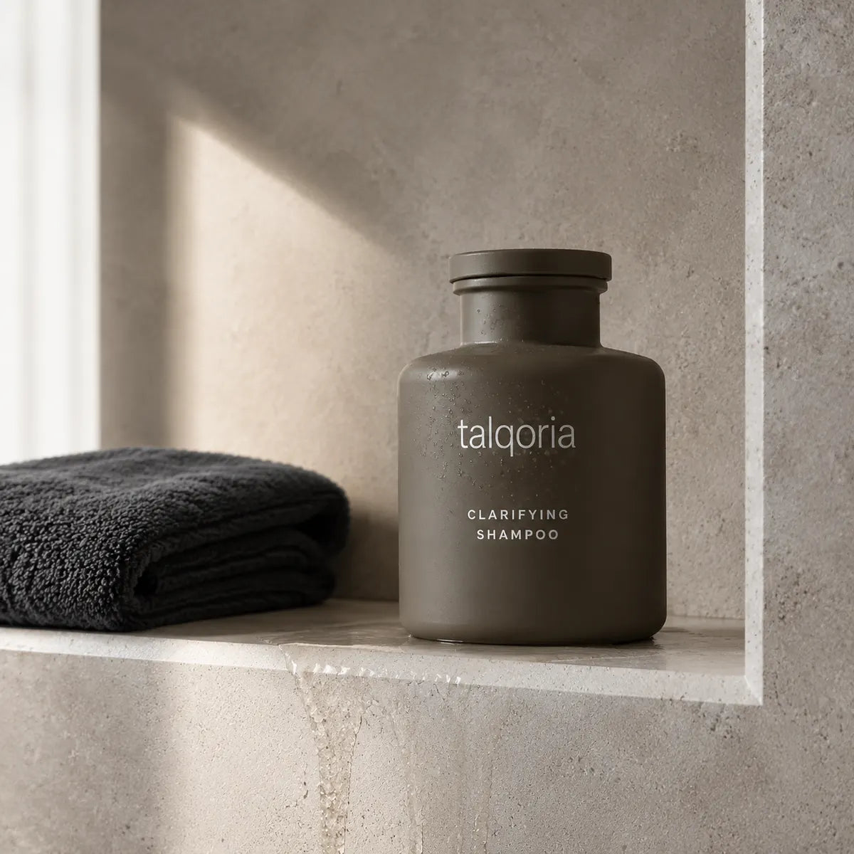 A matte dark gray bottle of talqoria Clarifying Shampoo sits on a stone shelf next to a folded dark gray towel, set against a textured stone wall with soft natural light.