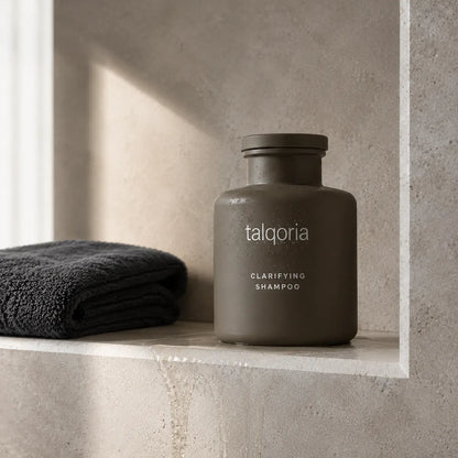 A matte dark gray bottle of talqoria Clarifying Shampoo sits on a stone shelf next to a folded dark gray towel, set against a textured stone wall with soft natural light.