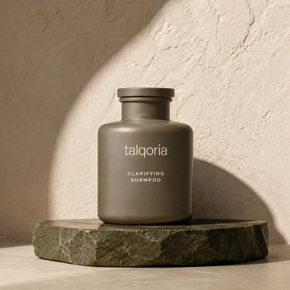 A matte taupe-gray bottle of talqoria Clarifying Shampoo sits on a dark stone slab against a textured beige wall with dramatic diagonal shadows.