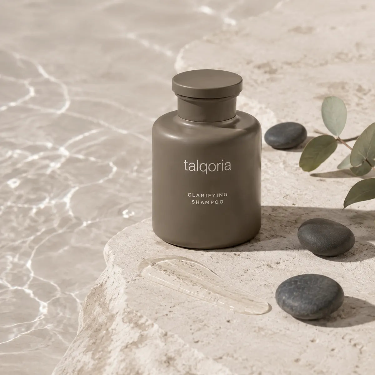 A matte taupe-gray bottle of talqoria Clarifying Shampoo sits on a textured stone slab next to smooth river stones and eucalyptus leaves, with a clear liquid smear in front of it.