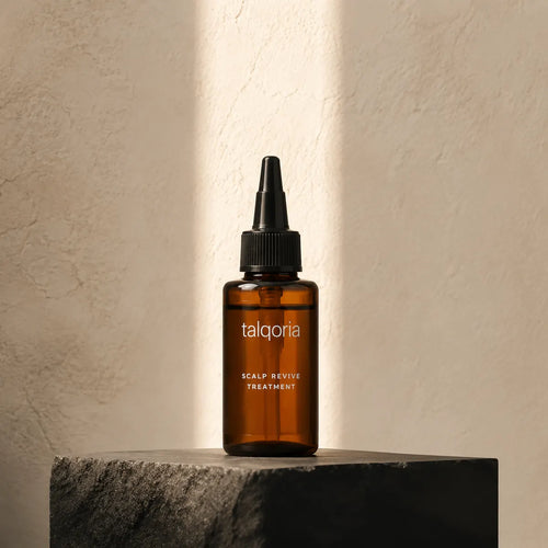 A small amber glass dropper bottle labeled talqoria Scalp Revive Treatment stands on a dark stone pedestal against a textured beige wall with warm sunlight and shadow streaks.