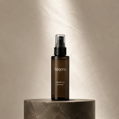 A dark amber glass spray bottle labeled 'talqoria LEAVE-IN ELIXIR' stands centered on a round stone pedestal against a softly lit beige wall.