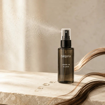 A dark amber glass spray bottle labeled 'talqoria LEAVE-IN ELIXIR' standing on a light stone surface with water droplets and strands of brown hair around it, against a softly lit beige background with mist rising from the spray.