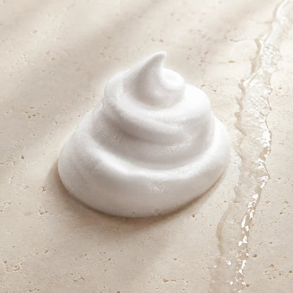 A dollop of thick, white whipped foam or shaving lather sits on a light beige textured surface with a glossy wet streak running along the right side.